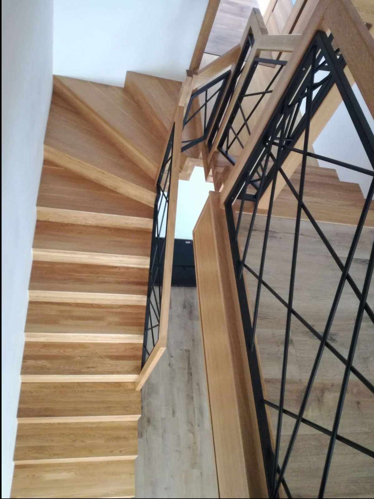 Drewniane schody z czarną, geometryczną balustradą, widok z góry. Jasne drewno stopni kontrastuje z ciemnym metalem balustrady.