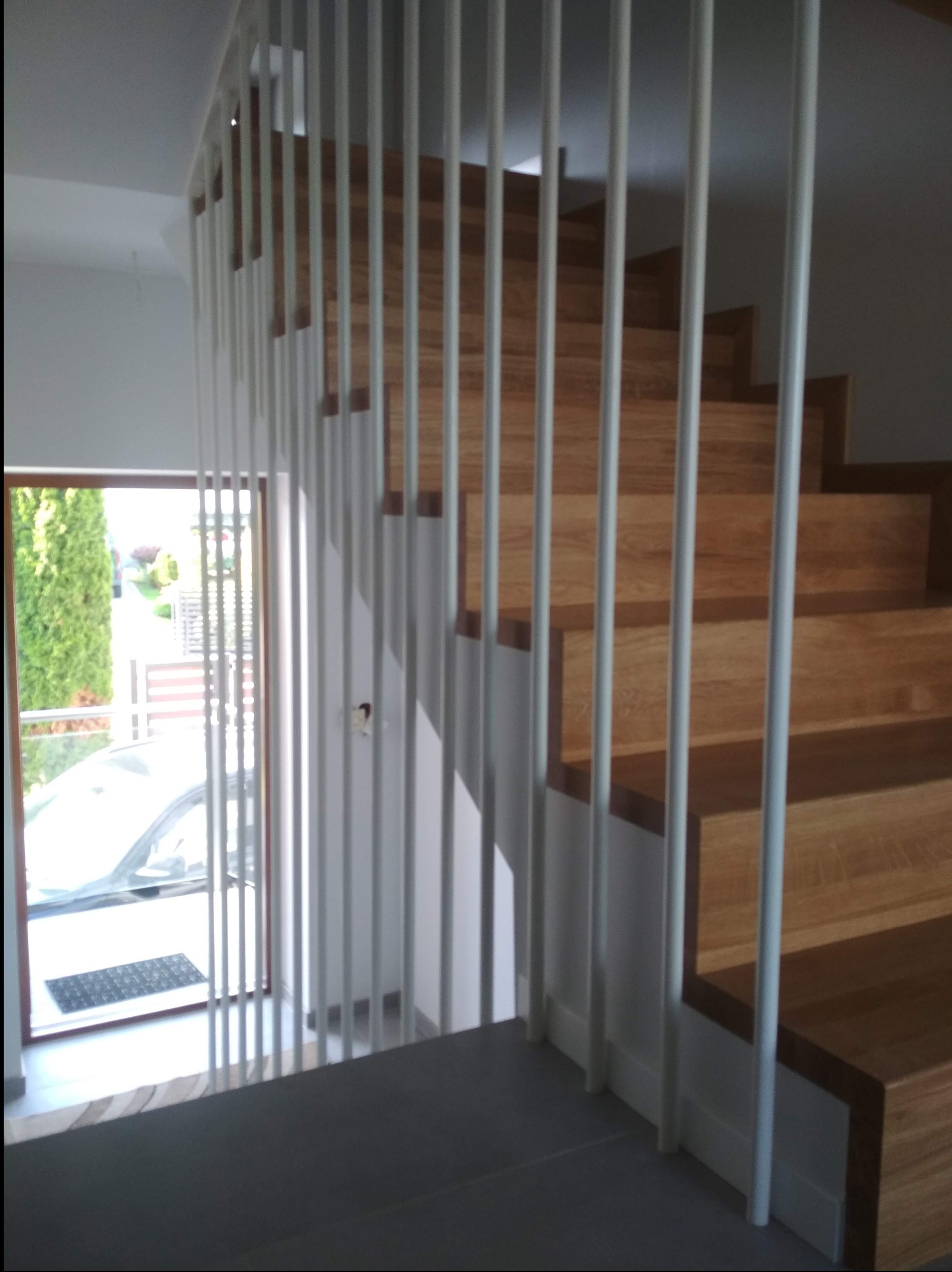 Nowoczesne schody z drewnianymi stopniami i białą, minimalistyczną balustradą z pionowych prętów. Widok z góry na jasne wnętrze z oknem i fragmentem otoczenia.
