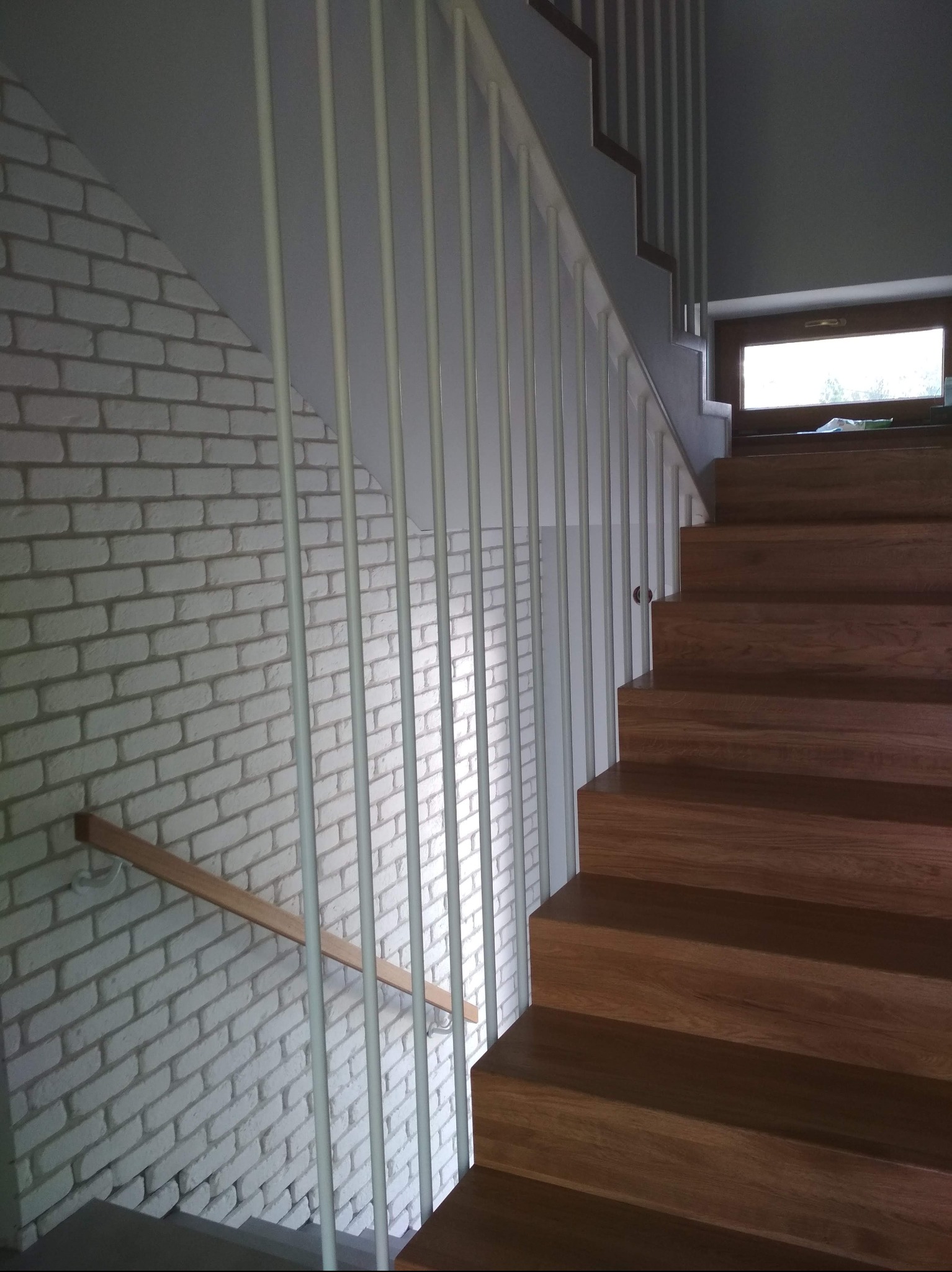 Drewniane schody z jasną balustradą na tle ściany z cegły. Widoczne stopnie i fragment okna. Ujęcie z dołu, podkreślające wysokość schodów.