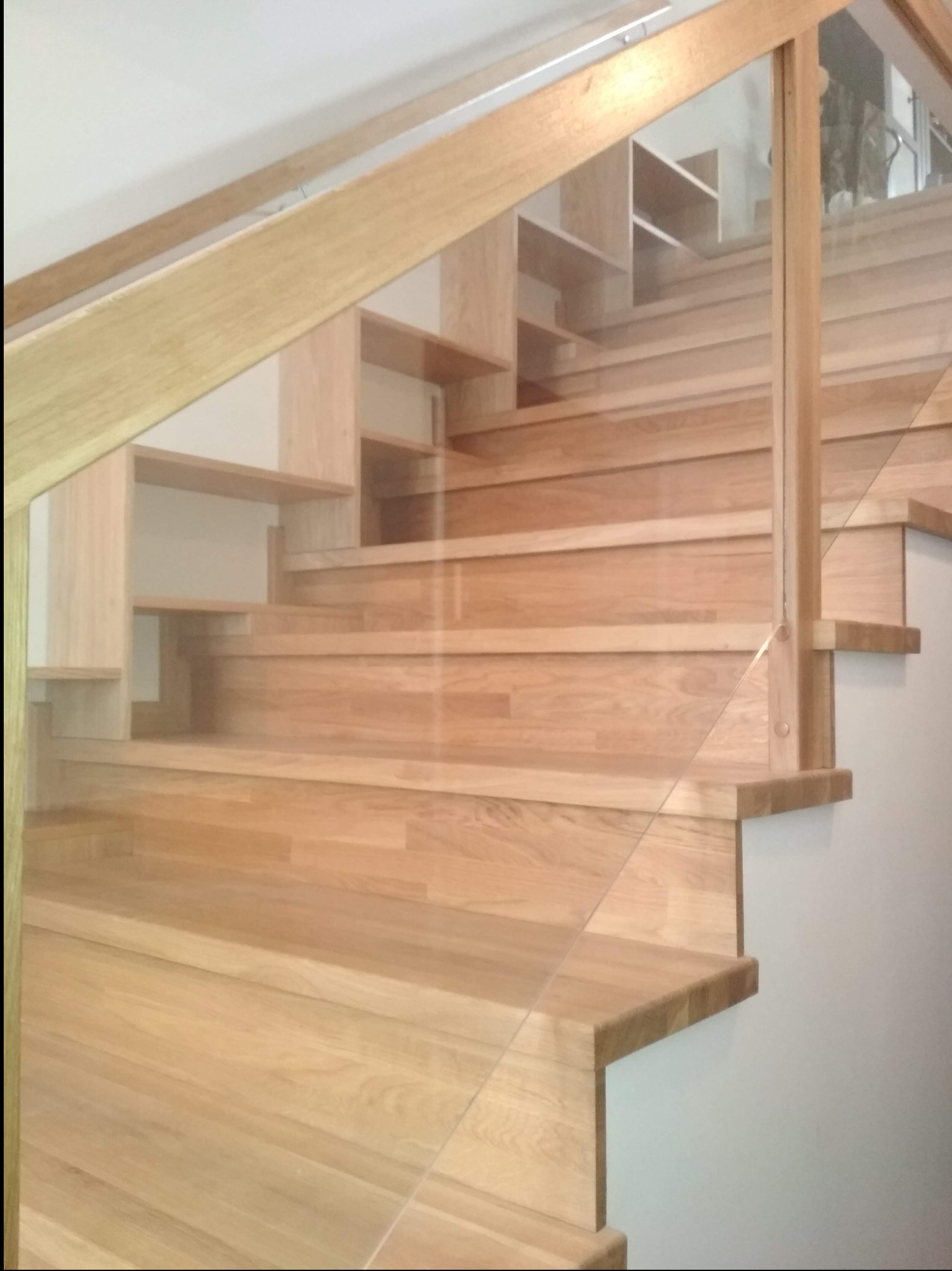 Jasne, drewniane schody z przeszkloną balustradą, widok z dołu. Stopnie zintegrowane z półkami, minimalistyczny design, naturalne oświetlenie.