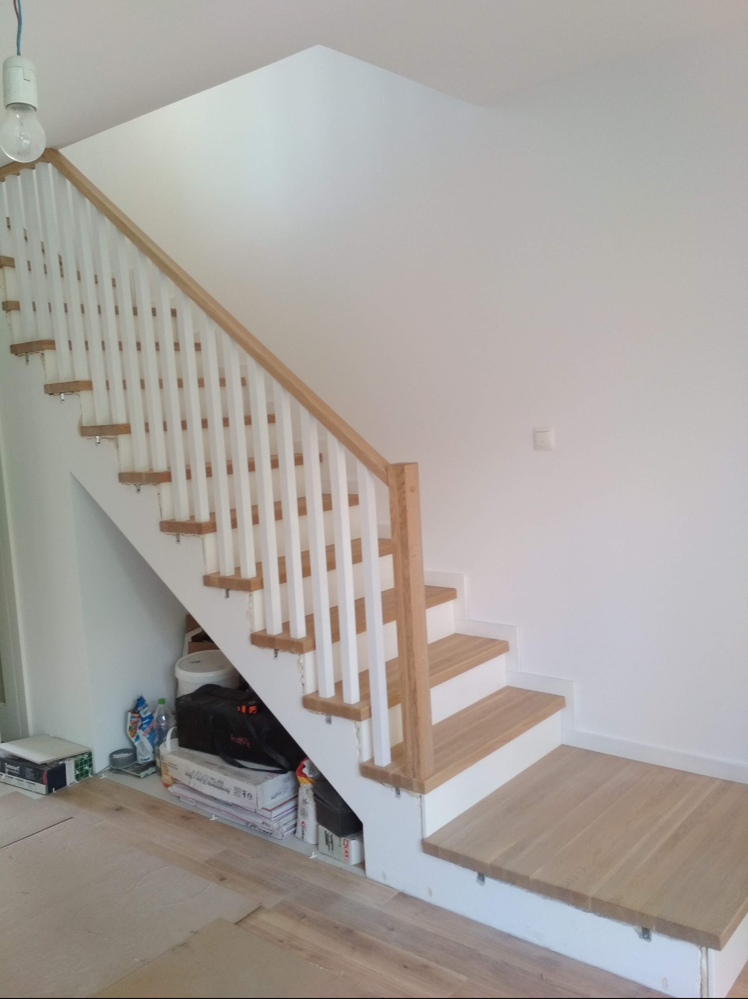 Jasne, drewniane schody z białymi balustradami w nowoczesnym wnętrzu. Widoczne detale wykończenia i oświetlenie. Częściowo widoczne materiały budowlane pod schodami.