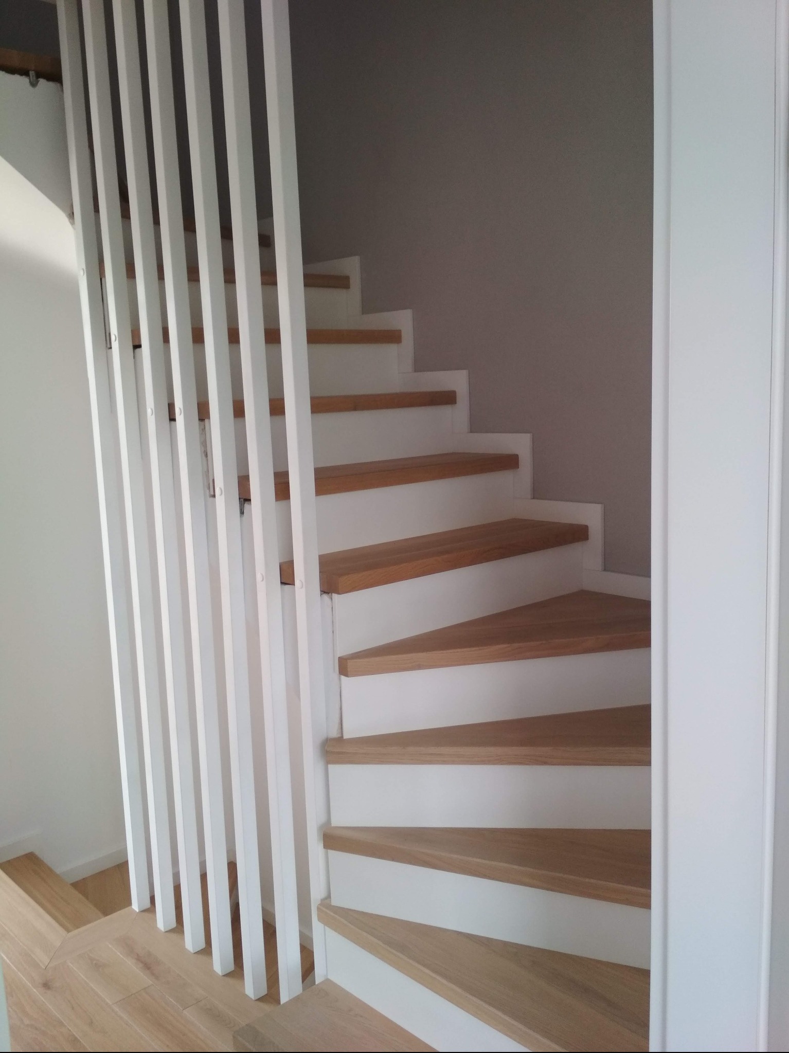 Biało-drewniane schody z ażurową barierką z pionowych listew, widoczne od dołu, z parkietem w jodełkę na podłodze. Minimalistyczny design, jasne wnętrze.