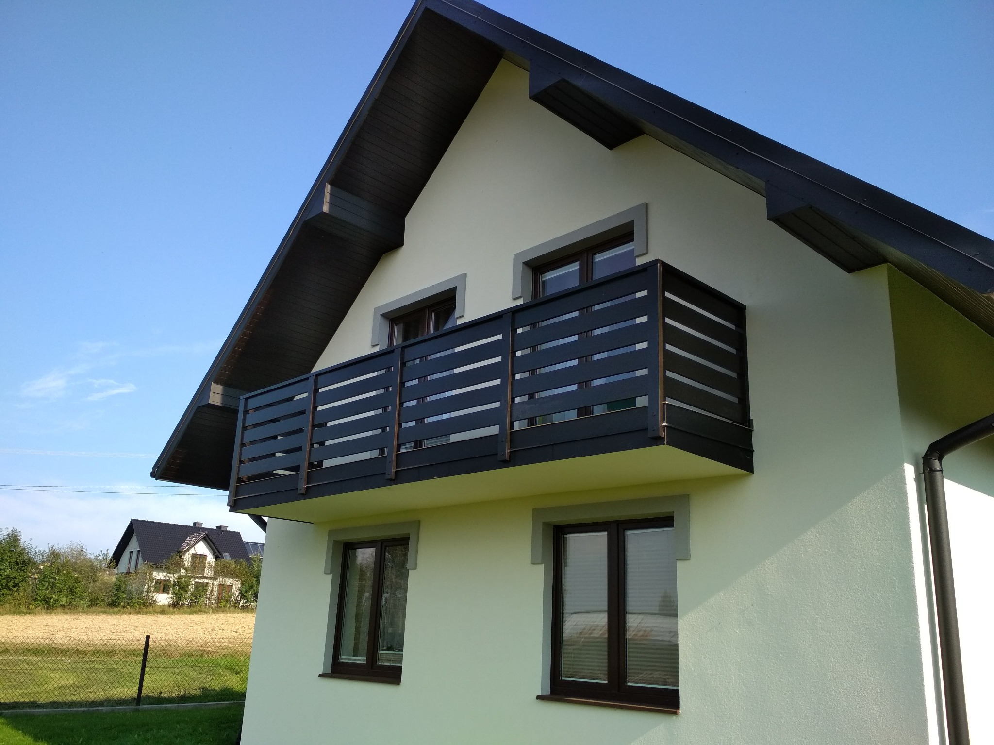 Elewacja domu z balkonem z czarną, nowoczesną balustradą. Widok na budynek z zewnątrz w słoneczny dzień. W tle pole i zabudowania.