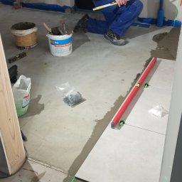 Oleg Andronik AndroBud - Układanie płytek podłogowych z użyciem poziomicy laserowej. Widoczny glazurnik, narzędzia i materiały budowlane. Prace w toku, surowy stan pomieszczenia.