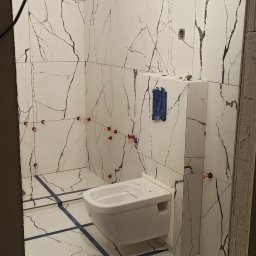 Oleg Andronik AndroBud - Biała łazienka w trakcie wykańczania: marmurowe płytki z czarnymi żyłami, niebieskie taśmy wyznaczające fugi i pomarańczowe kliny poziomujące płytki. Podwieszana toaleta.