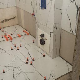 Oleg Andronik AndroBud - Białe płytki imitujące marmur na ścianach i podłodze łazienki w trakcie montażu. Widoczne poziomice i system poziomowania płytek.