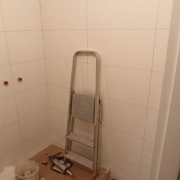 Oleg Andronik AndroBud - Remont łazienki w trakcie: białe płytki ścienne, wzorzysta podłoga, drabina i narzędzia glazurnicze. Widoczne przygotowane otwory na armaturę.