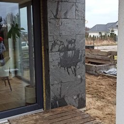 Oleg Andronik AndroBud - Wykończona ściana zewnętrzna z szarej, kamiennej okładziny na balkonie. Widoczna paleta i elementy konstrukcyjne. W tle domy.