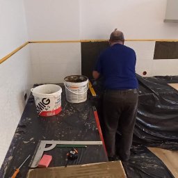 Oleg Andronik AndroBud - Glazurnik w trakcie pracy: układanie białych płytek na ścianie w kuchni. Widoczne narzędzia, wiaderka z klejem i folia ochronna. Częściowo ułożone płytki.
