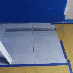 Oleg Andronik AndroBud - Ułożone szare płytki podłogowe w łazience z odpływem liniowym. Ściany częściowo pomalowane na niebiesko, widoczne narzędzia i wiadro z zaprawą. Prace w toku.