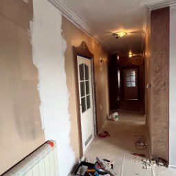 Oleg Andronik AndroBud - Korytarz w trakcie malowania: fragment ściany pomalowany na biało, zabezpieczona podłoga, widoczne narzędzia malarskie i grzejnik. Dekoracyjne wykończenie sufitu.