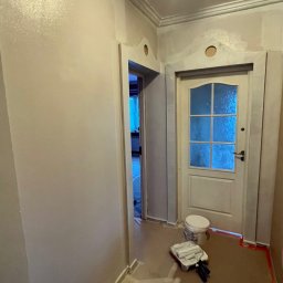 Oleg Andronik AndroBud - Korytarz w trakcie malowania na szaro, widoczne wałki malarskie, wiadro z farbą, zabezpieczona podłoga i świeżo pomalowane ściany z dekoracyjną listwą pod sufitem.