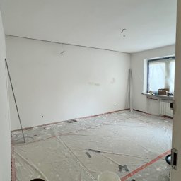 Oleg Andronik AndroBud - Pokój w trakcie przygotowań do malowania: zabezpieczona podłoga folią, widoczne puszki z farbą, świeżo pomalowane ściany na biało, okno z grzejnikiem.