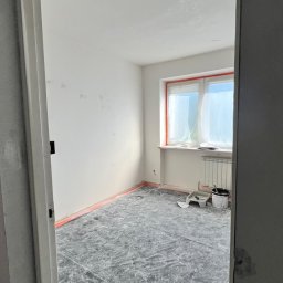 Oleg Andronik AndroBud - Pokój w trakcie malowania: zabezpieczona podłoga, okno z folią, ściany po szpachlowaniu, widoczne narzędzia malarskie. Ujęcie zza futryny.