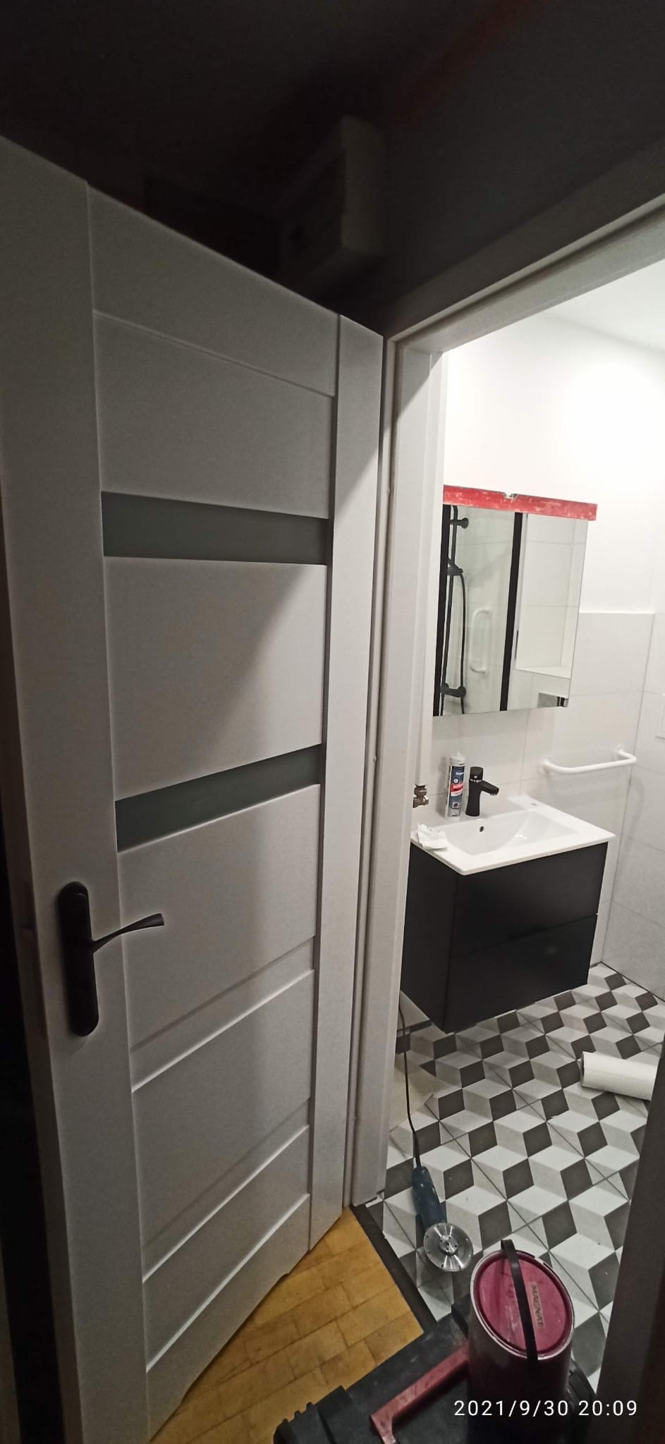 Białe drzwi z poziomymi przeszkleniami, prowadzące do wykończonej łazienki z czarno-białą podłogą i czarną szafką pod umywalką. Widoczne narzędzia i farba.