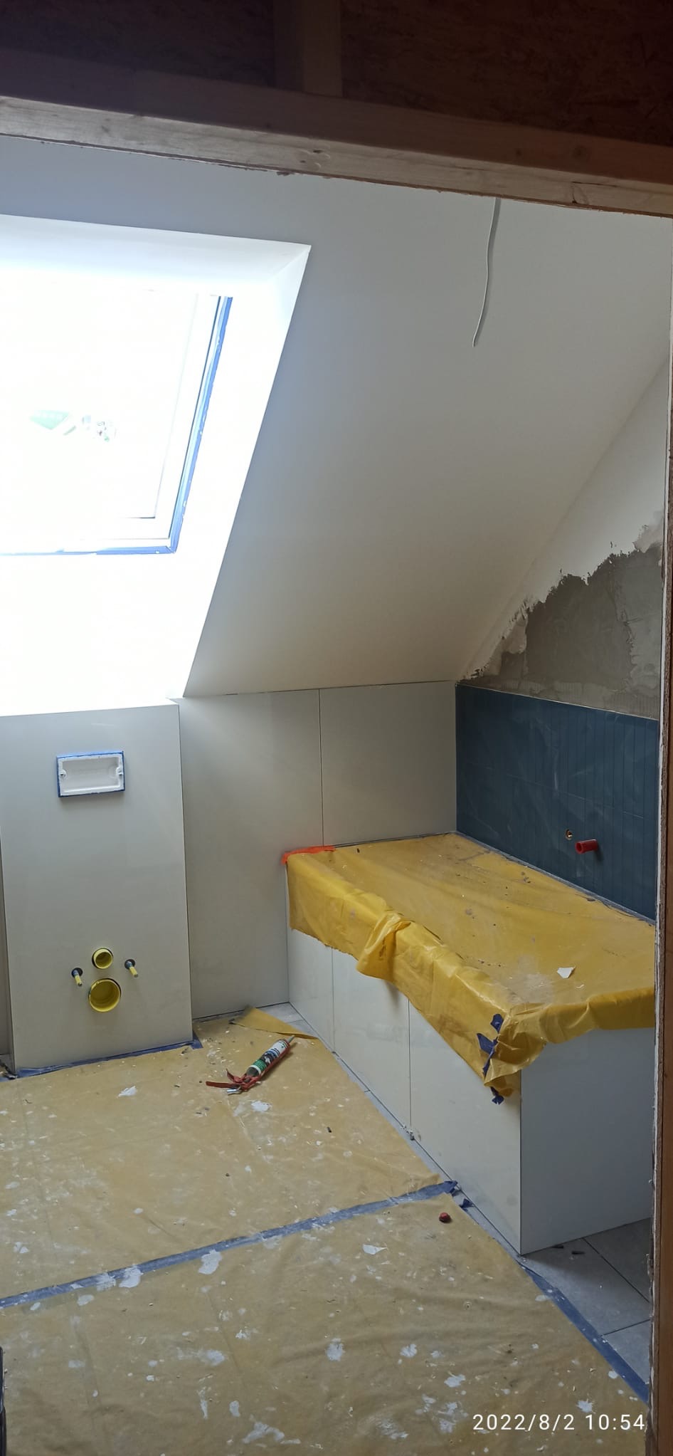 Łazienka w trakcie wykańczania: białe płytki na ścianach, niebieska mozaika, okno dachowe. Podłoga zabezpieczona folią, widoczne narzędzia i elementy instalacji.