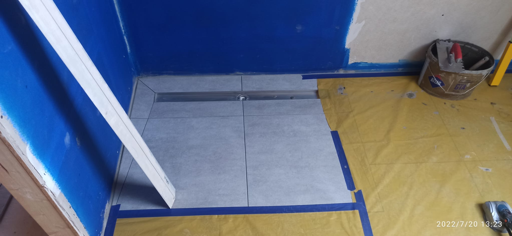 Ułożone szare płytki podłogowe w łazience z odpływem liniowym. Ściany częściowo pomalowane na niebiesko, widoczne narzędzia i wiadro z zaprawą. Prace w toku.