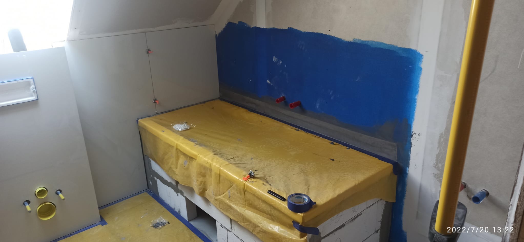 Łazienka w trakcie prac: zabezpieczona folią wanna, ściany z niebieską hydroizolacją i częściowo położonymi płytkami. Widoczne narzędzia i elementy instalacji.