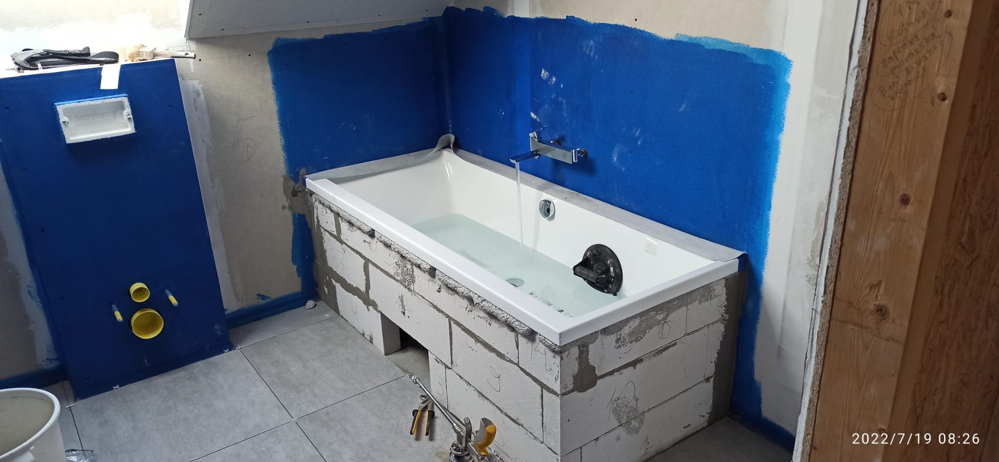 Wanna obudowana bloczkami, ściany zabezpieczone folią w płynie. Widoczne narzędzia i fragment instalacji. Etap prac wykończeniowych w łazience.