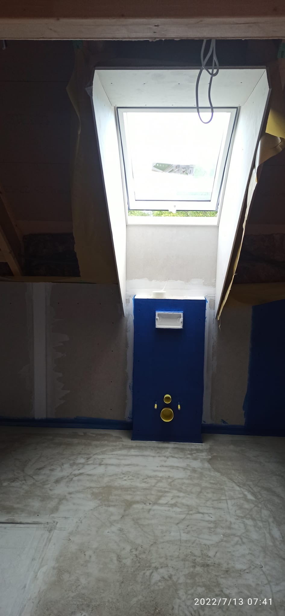 Niebieska zabudowa stelaża WC pod oknem dachowym na poddaszu w trakcie prac wykończeniowych. Widoczne przygotowane instalacje. Surowy beton na podłodze.