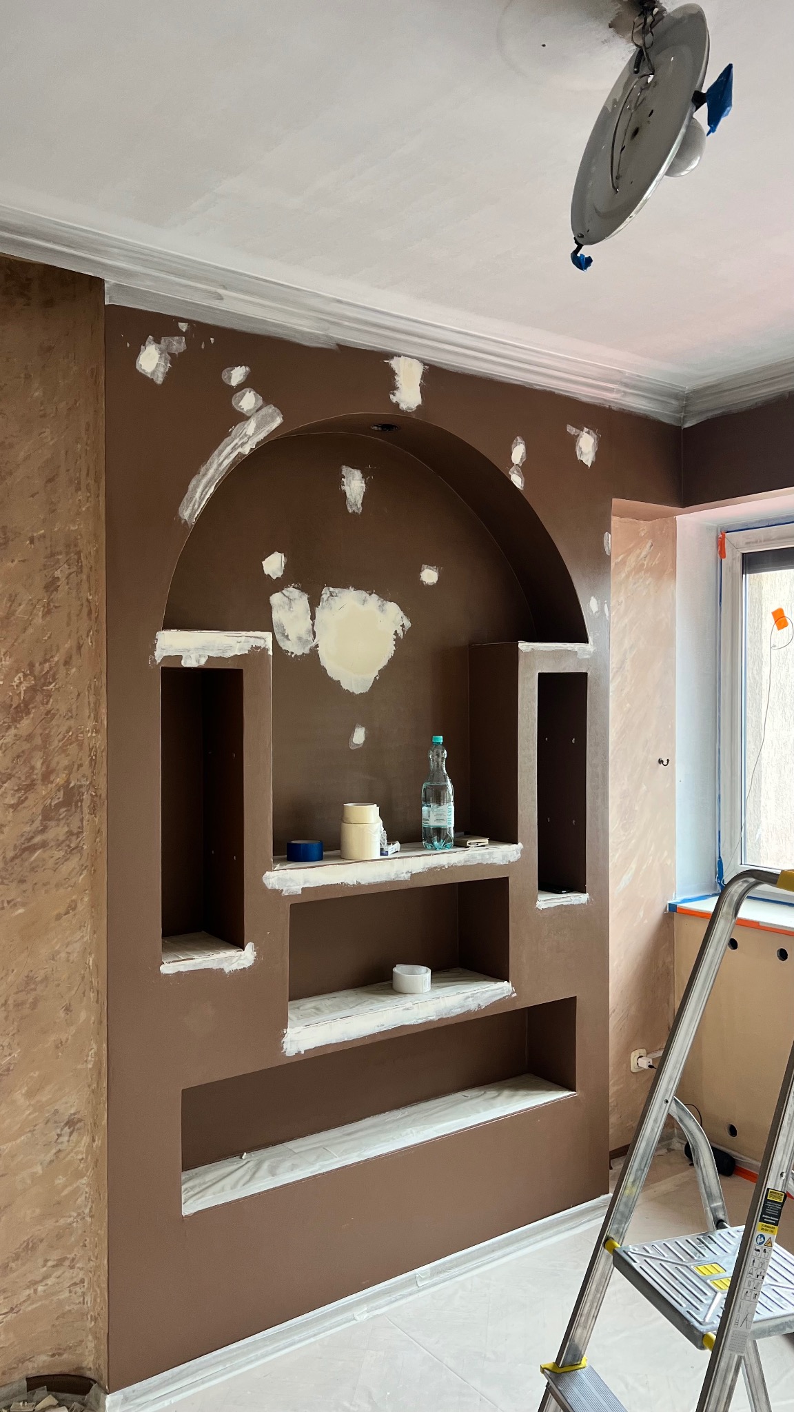 Wnęka w ścianie w trakcie szpachlowania i malowania na brązowo. Widoczne ślady po szpachli, drabina i akcesoria malarskie. Etap prac wykończeniowych.