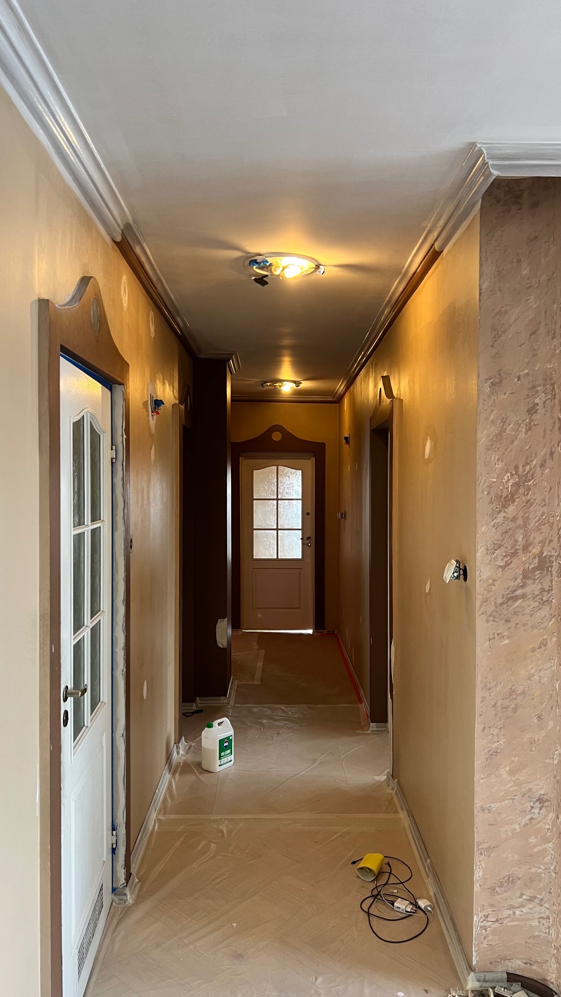 Korytarz w trakcie prac wykończeniowych: zabezpieczona podłoga folią, ściany po szpachlowaniu z efektem dekoracyjnym, widoczne drzwi z ościeżnicami i listwy sufitowe.