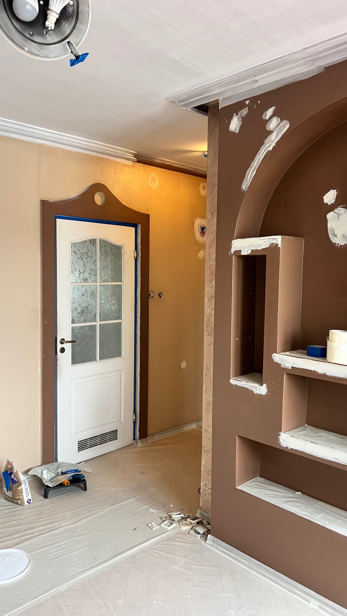 Wnętrze w trakcie prac wykończeniowych: brązowa ściana z wnękami, białe elementy dekoracyjne, zabezpieczona podłoga, widoczne narzędzia malarskie i puszki z farbą.