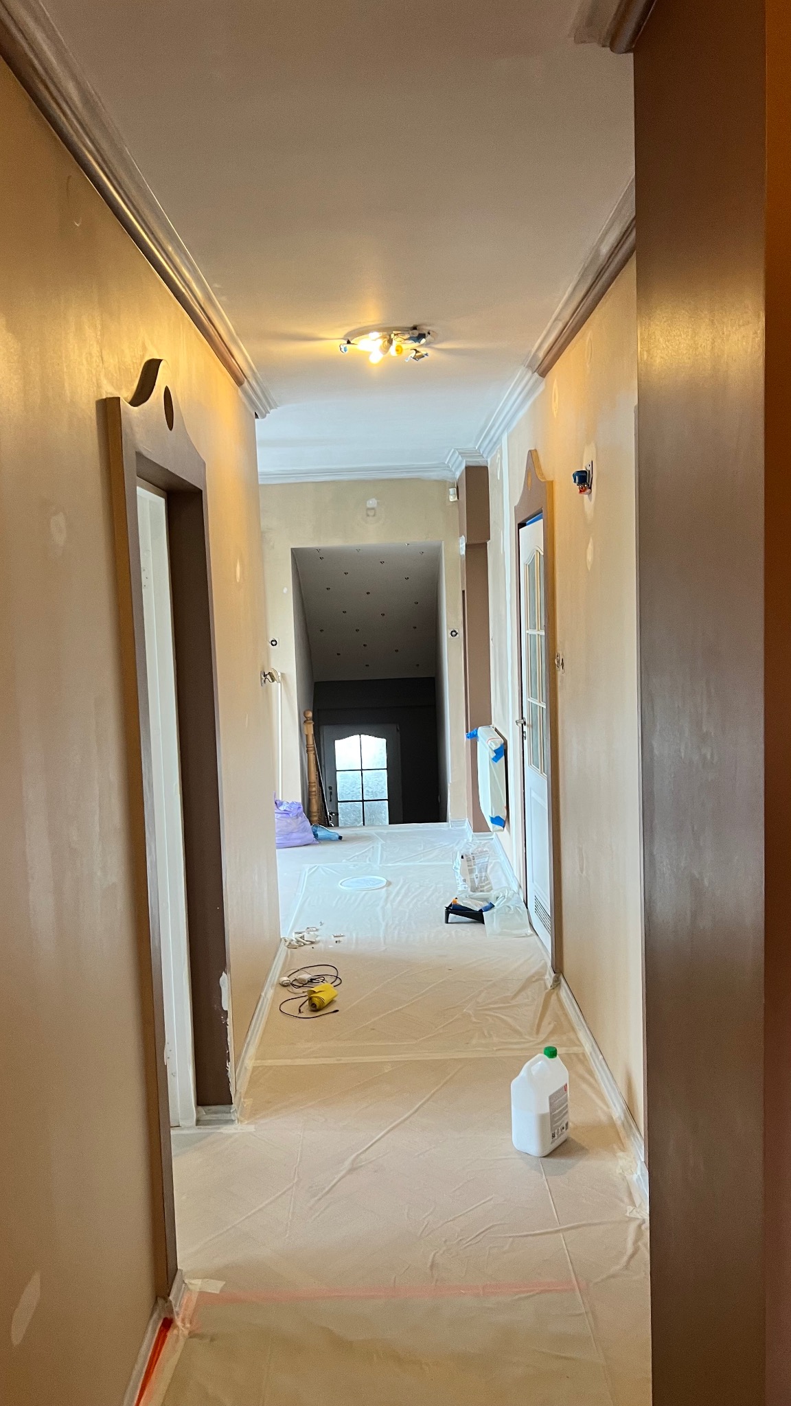 Korytarz w trakcie prac wykończeniowych: ściany po szpachlowaniu, zabezpieczona podłoga, widoczne narzędzia i materiały malarskie. Przygotowanie do malowania.