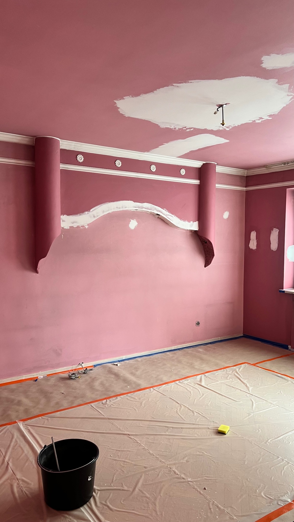 Różowe wnętrze w trakcie prac wykończeniowych: ściany z dekoracyjnymi elementami, częściowo wyszpachlowane, podłoga zabezpieczona folią malarską, widoczne narzędzia.