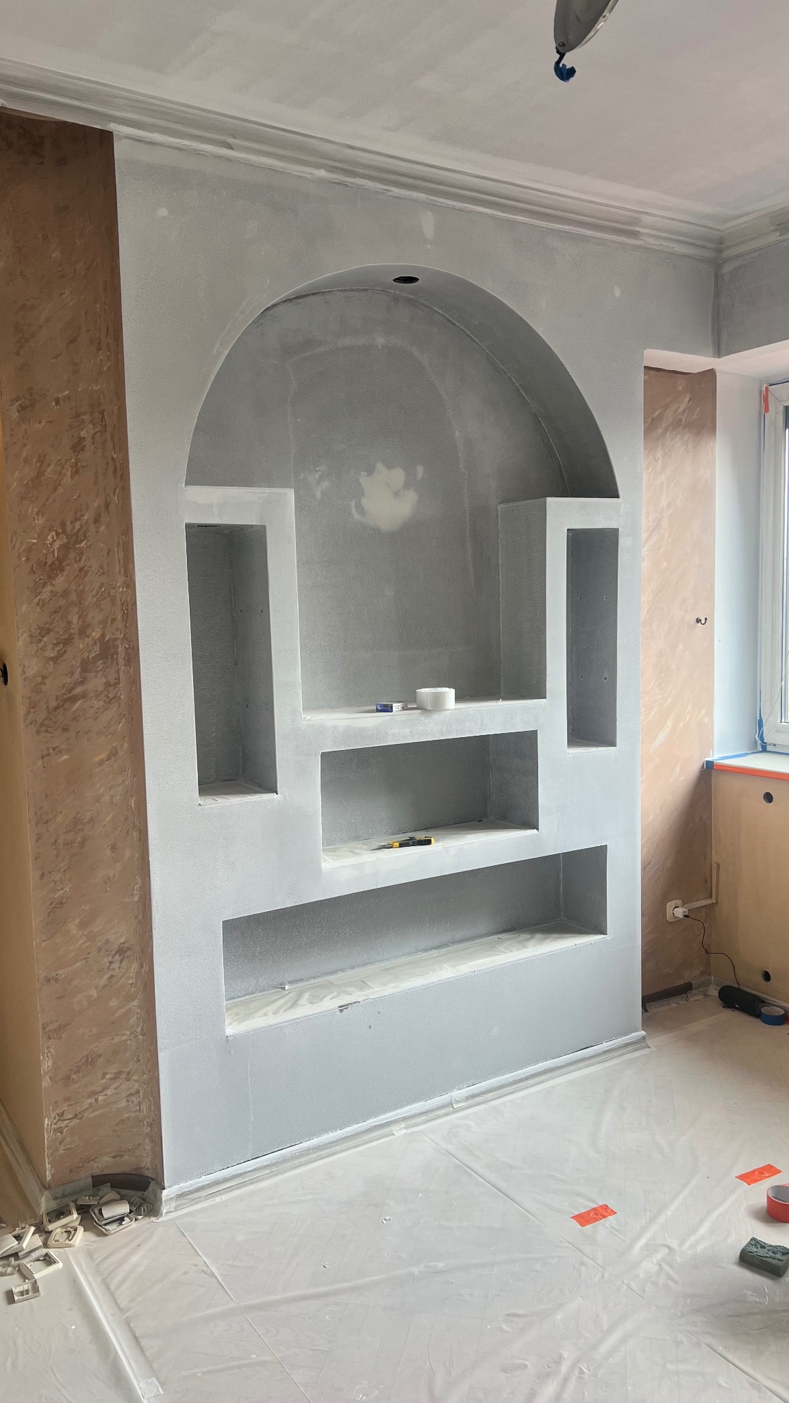 Narożna wnęka z półkami w kształcie łuku, świeżo wyszpachlowana na szaro, z widocznymi narzędziami i folią ochronną na podłodze. Ściany częściowo przygotowane do malowania.