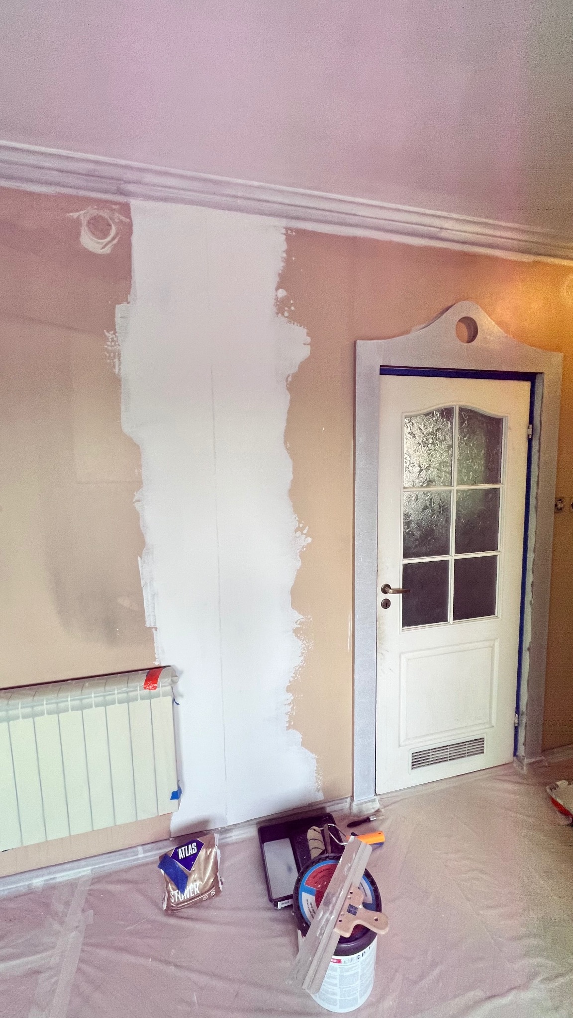 Ściana w trakcie szpachlowania, widoczny fragment grzejnika, drzwi z ościeżnicą i narzędzia malarskie na podłodze zabezpieczonej folią. Częściowo nałożona biała gładź.