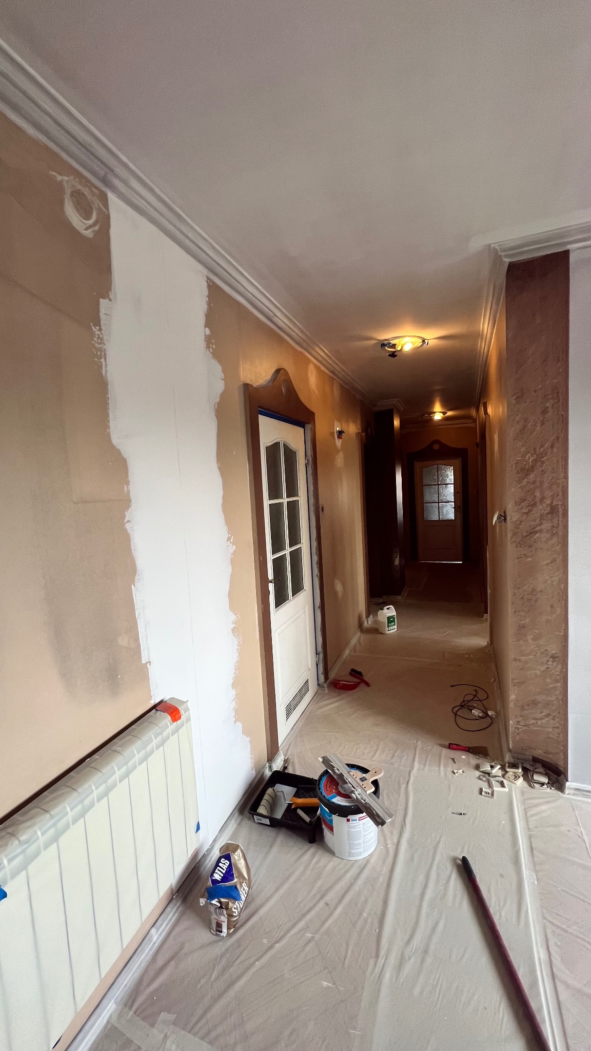 Korytarz w trakcie malowania: fragment ściany pomalowany na biało, zabezpieczona podłoga, widoczne narzędzia malarskie i grzejnik. Dekoracyjne wykończenie sufitu.