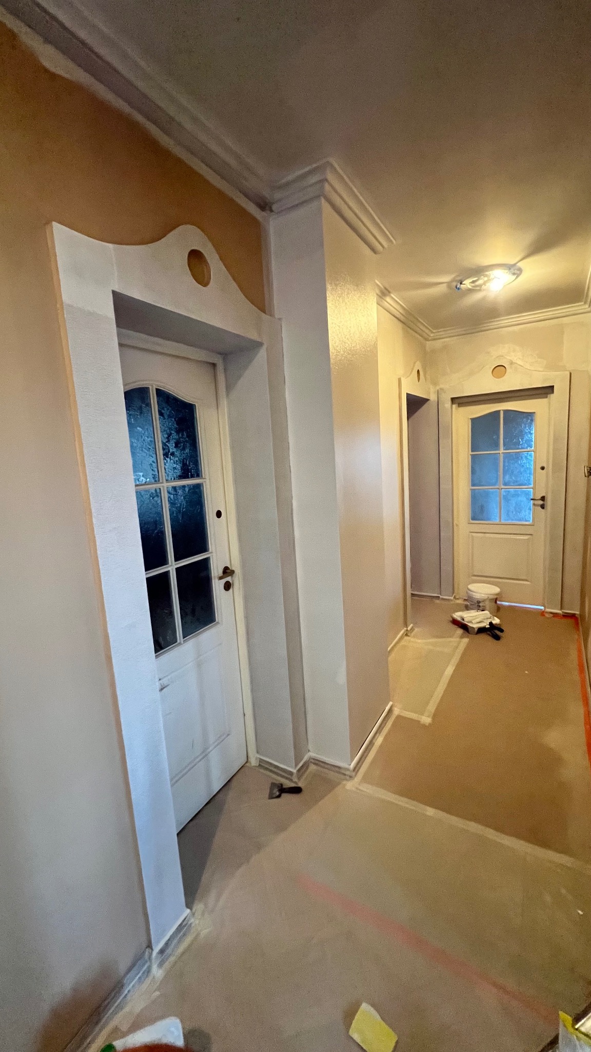 Korytarz w trakcie malowania: ściany pokryte świeżą warstwą farby, zabezpieczona podłoga, widoczne narzędzia malarskie i charakterystyczne obramowania drzwi.