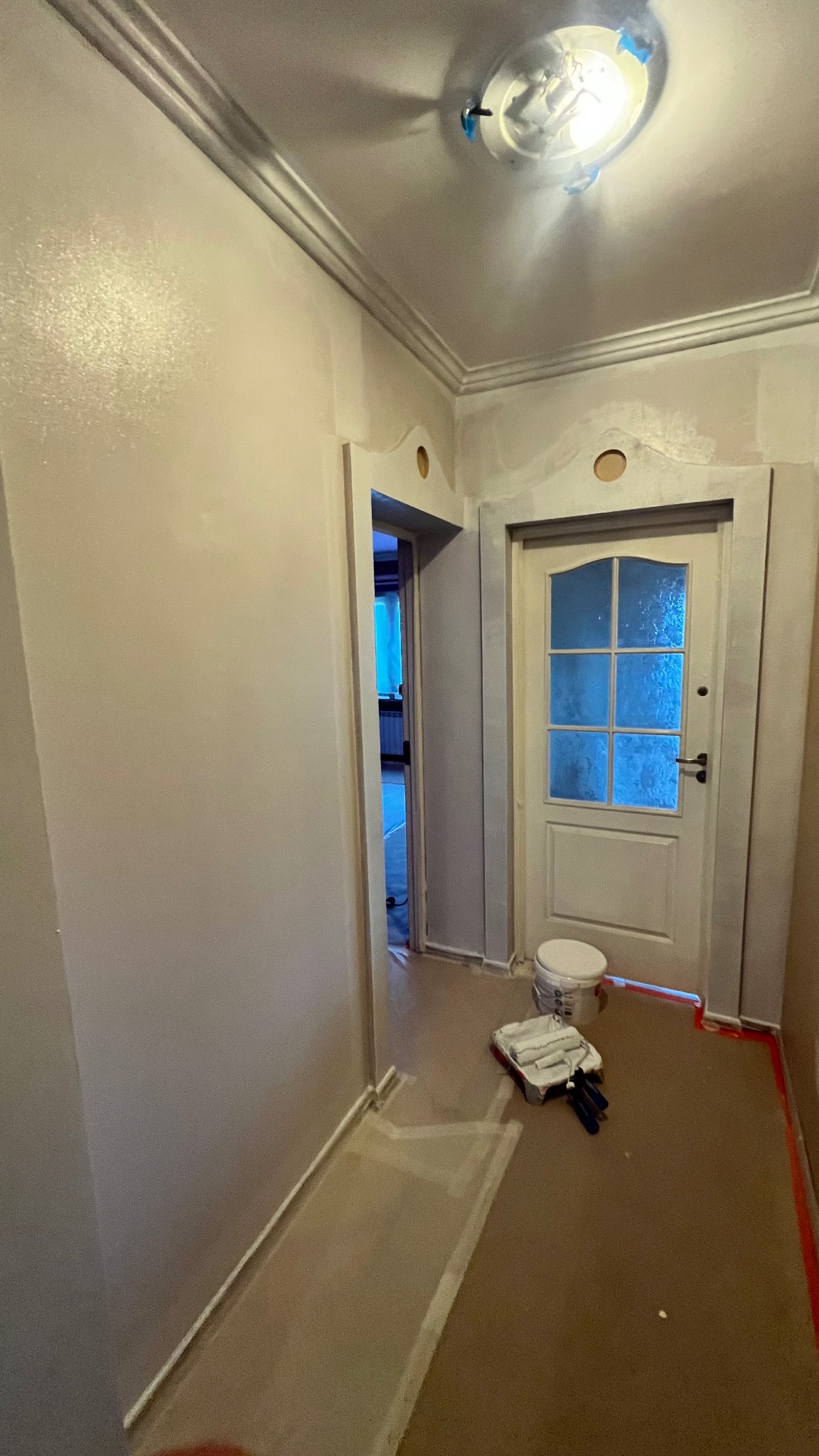 Korytarz w trakcie malowania na szaro, widoczne wałki malarskie, wiadro z farbą, zabezpieczona podłoga i świeżo pomalowane ściany z dekoracyjną listwą pod sufitem.