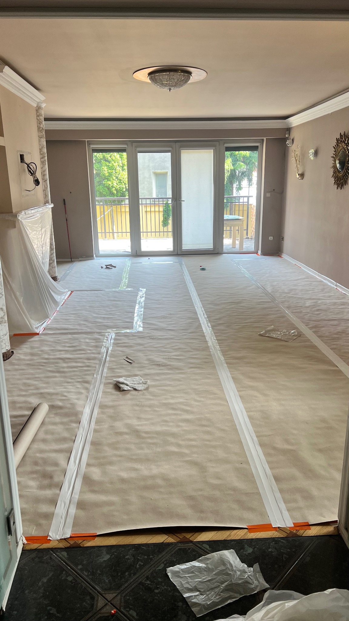 Pokój przygotowany do malowania: podłoga zabezpieczona papierem i taśmą malarską, widoczne okna balkonowe i elementy dekoracyjne na ścianach. Przygotowanie do prac malarskich.