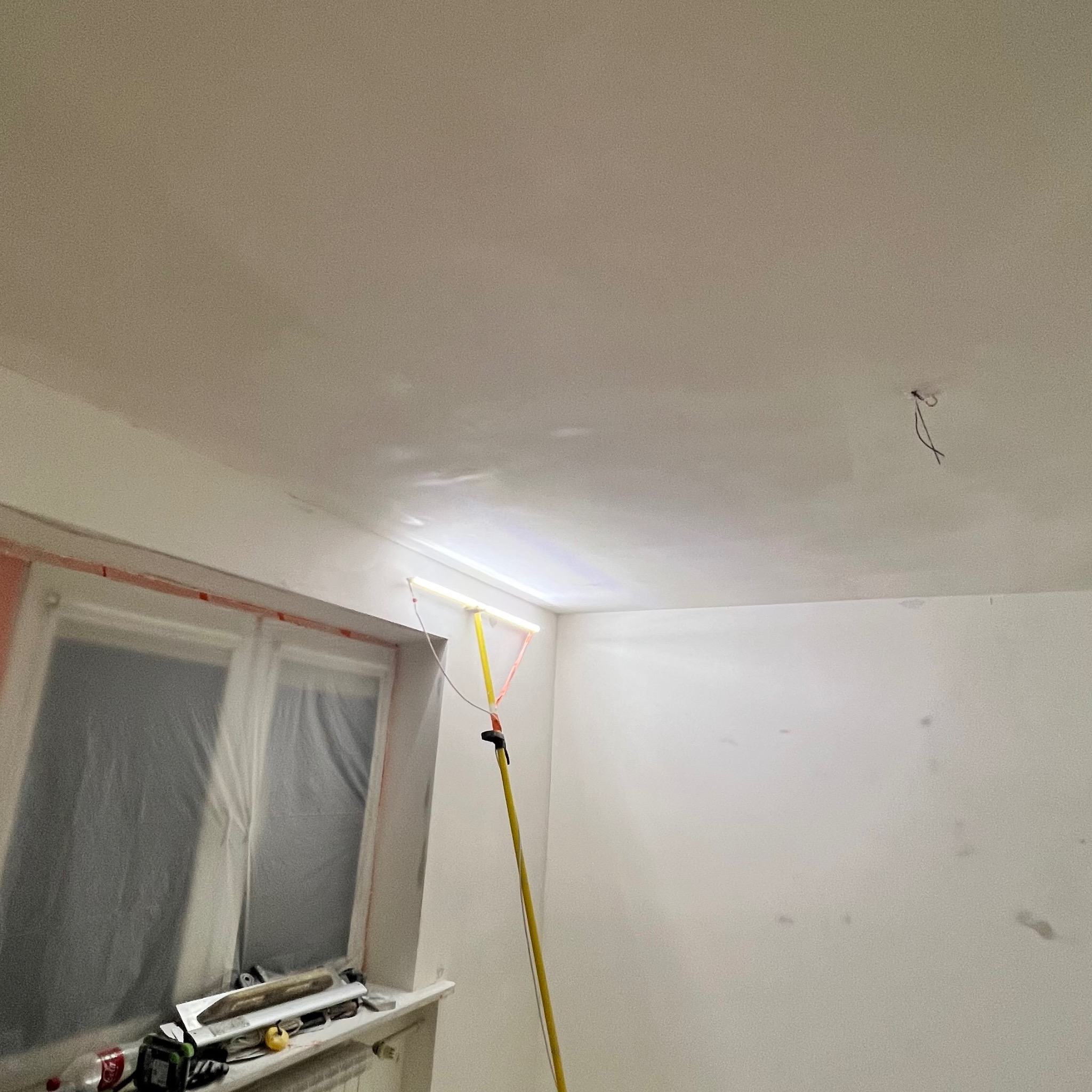Pokój w trakcie malowania: ściany i sufit po szpachlowaniu, okno zabezpieczone folią, lampa inspekcyjna na wysięgniku oświetla narożnik. Widoczne narzędzia na parapecie.
