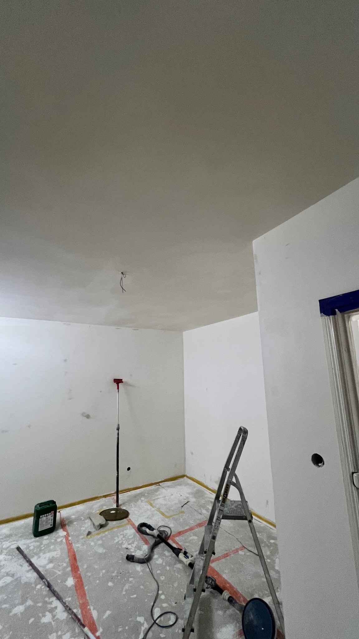 Pokój w trakcie szpachlowania: białe ściany z widocznymi śladami szpachli, drabina, szlifierka do gipsu, taśmy malarskie i narzędzia. Widok z dołu.