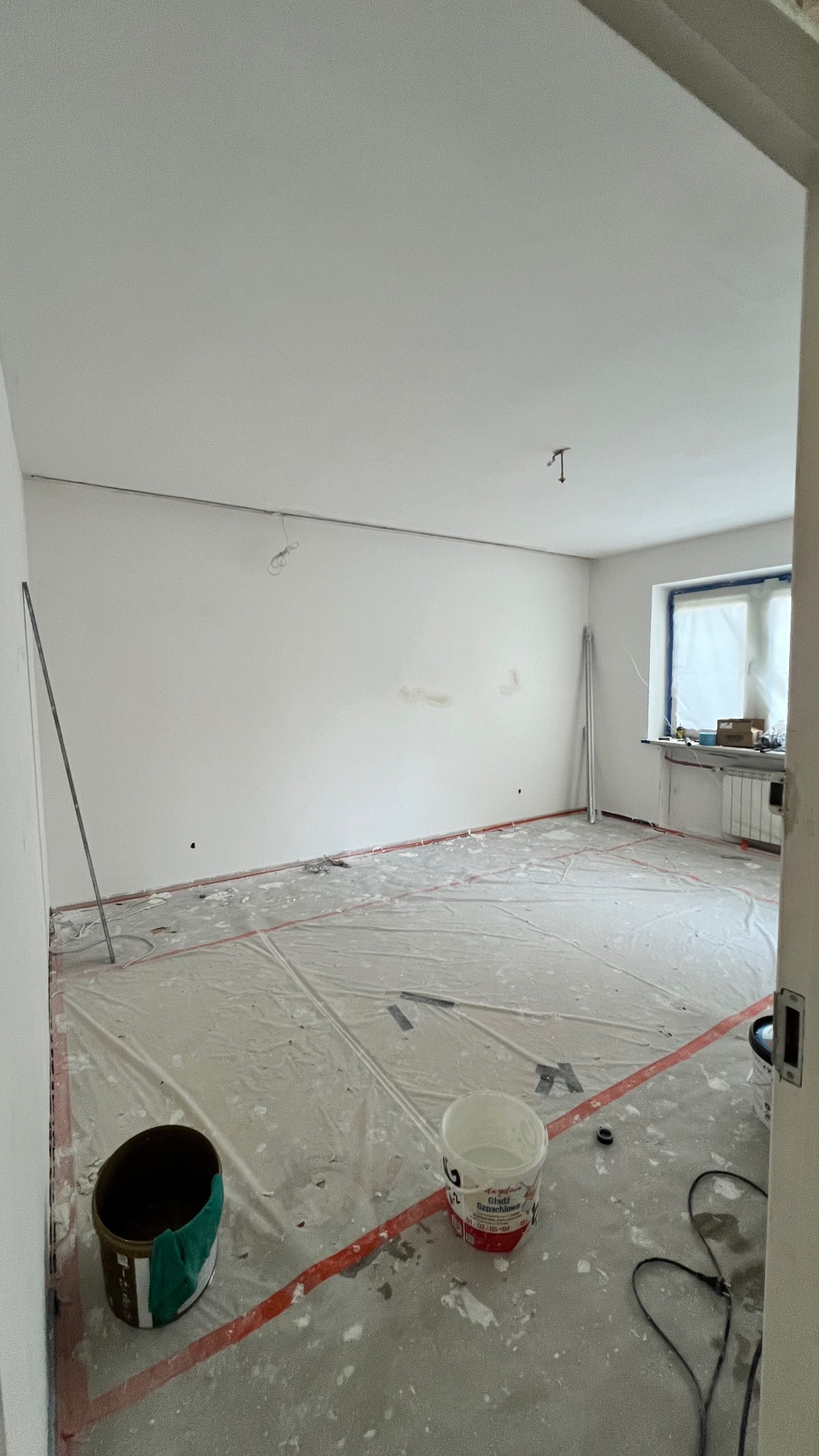 Pokój w trakcie przygotowań do malowania: zabezpieczona podłoga folią, widoczne puszki z farbą, świeżo pomalowane ściany na biało, okno z grzejnikiem.