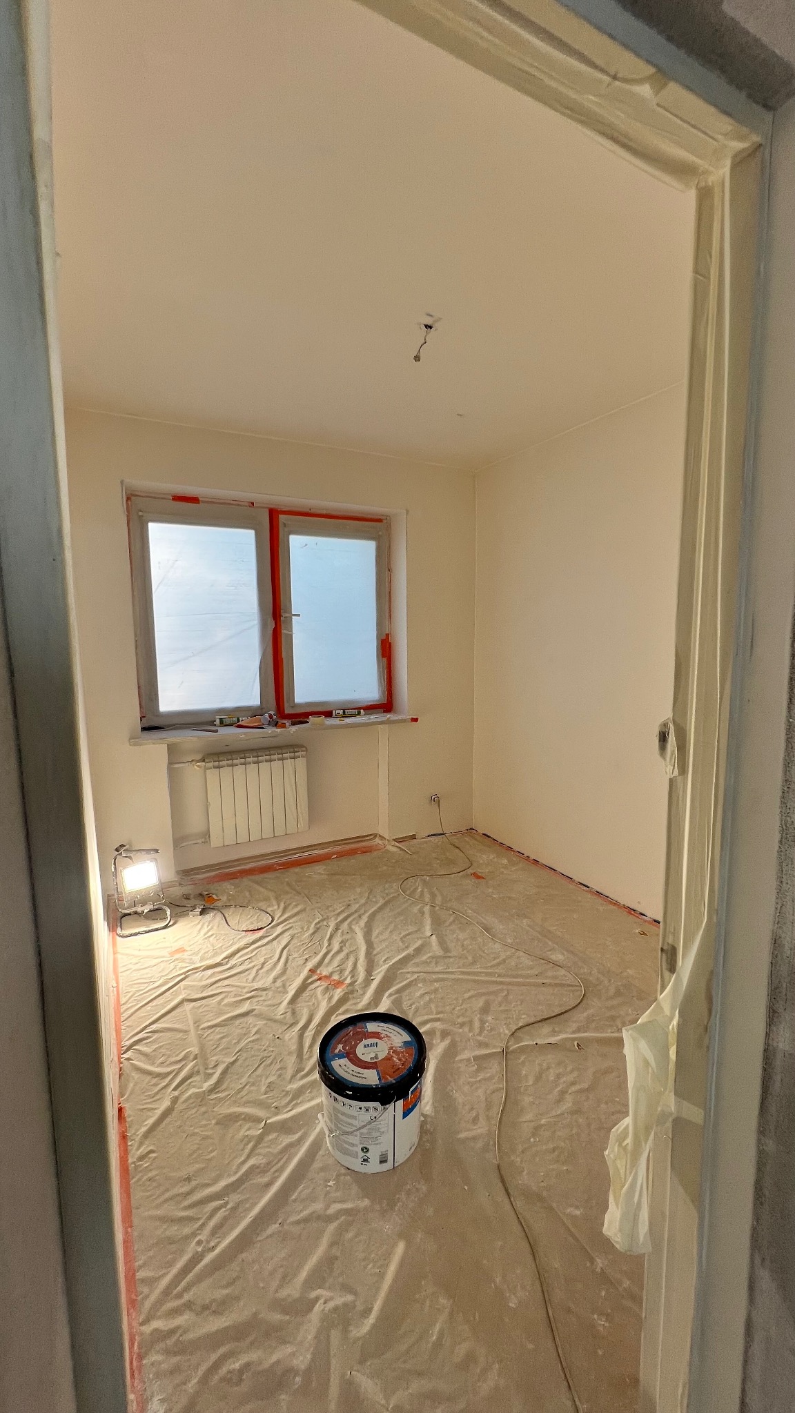 Pokój po szpachlowaniu, przygotowany do malowania. Okna i grzejnik zabezpieczone folią, podłoga przykryta, widoczna puszka z farbą i lampa oświetlająca.