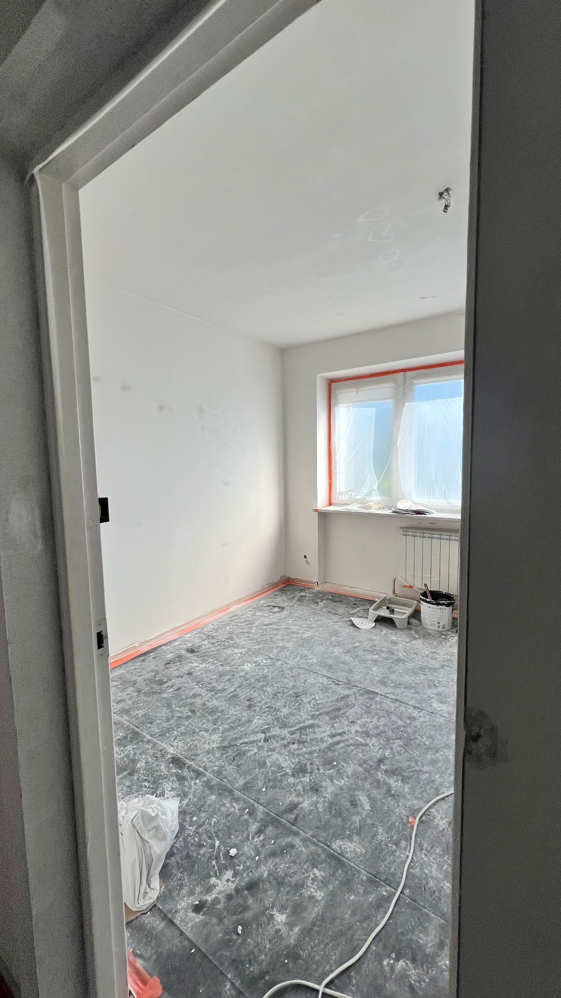 Pokój w trakcie malowania: zabezpieczona podłoga, okno z folią, ściany po szpachlowaniu, widoczne narzędzia malarskie. Ujęcie zza futryny.