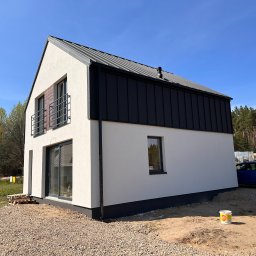 Steelvio House - Nowoczesny dom z białymi ścianami i czarnym dachem z blachy na tle błękitnego nieba. Widoczne okna, balkon z metalową balustradą i teren wokół domu w trakcie prac wykończeniowych.