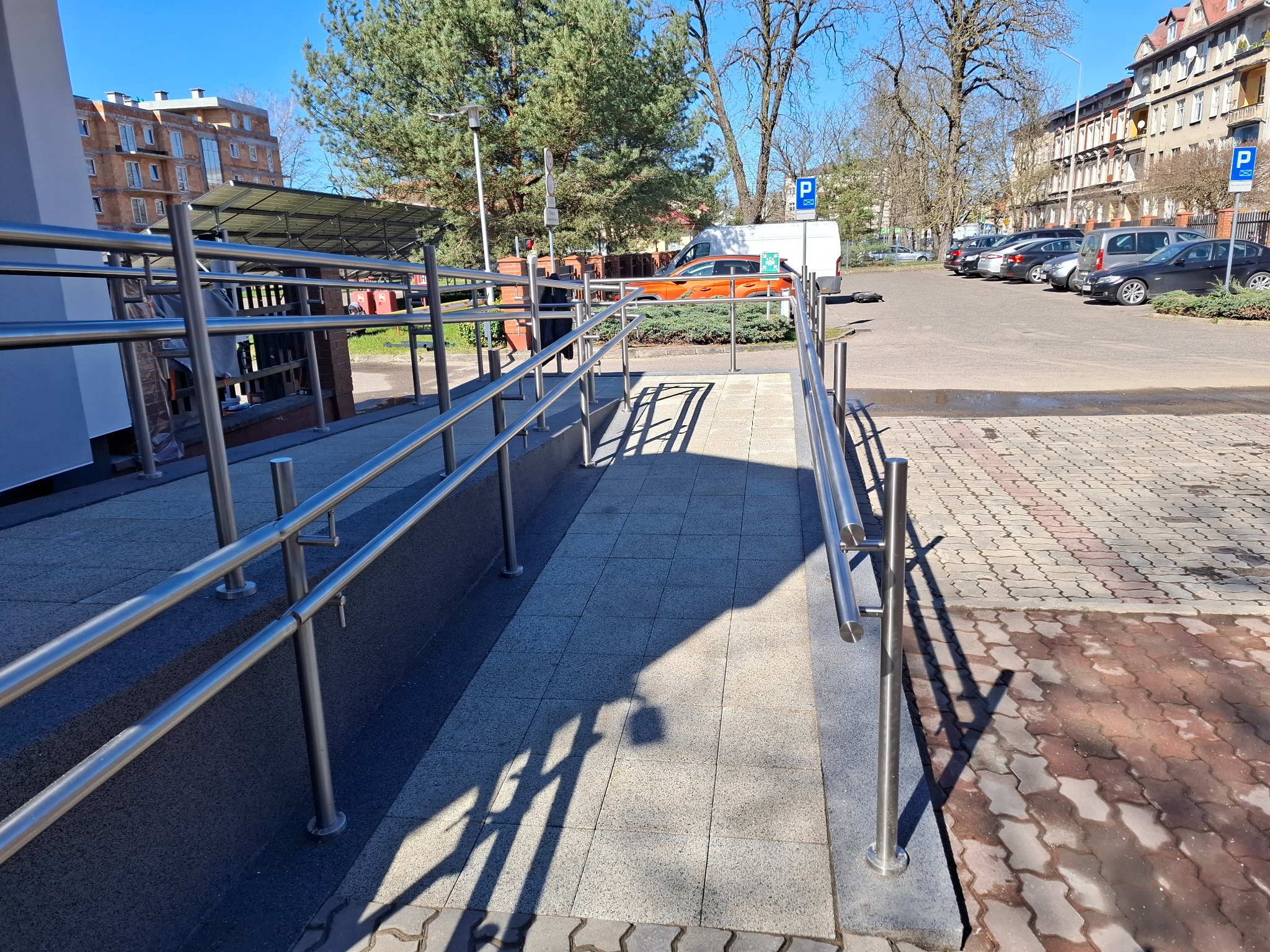 Zewnętrzna rampa dla niepełnosprawnych z metalowymi poręczami, wyłożona szarą kostką brukową, prowadząca do budynku. W tle parking i zabudowa miejska w słoneczny dzień.