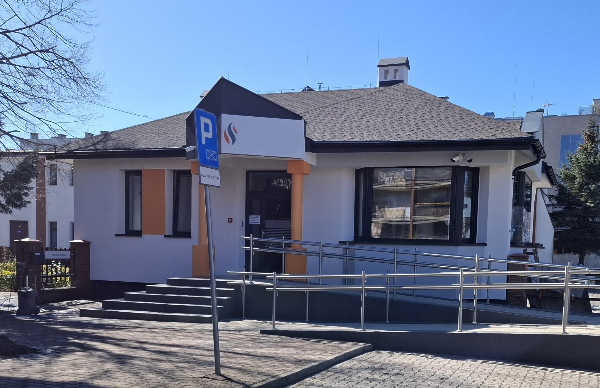 Biały budynek biurowy z rampą dla niepełnosprawnych, pomalowany na biało z pomarańczowymi akcentami, widoczny znak parkingu dla klientów i logo firmy.