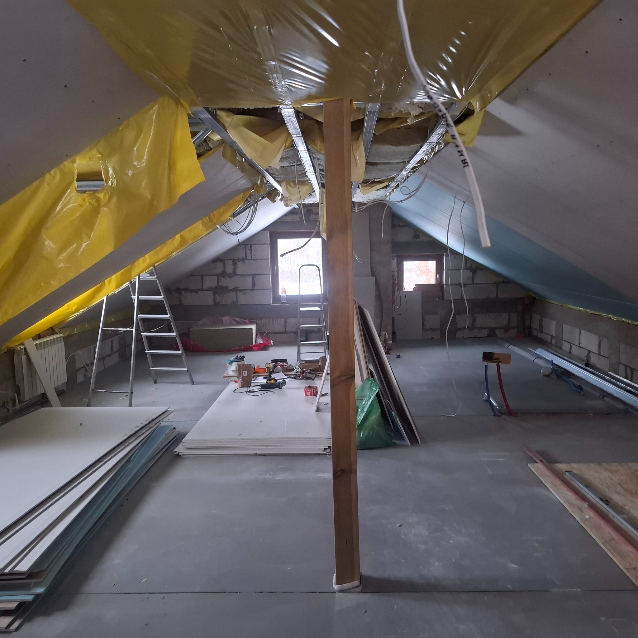 Poddasze w trakcie adaptacji: widoczne płyty karton-gips, metalowa konstrukcja sufitu, izolacja folią, okna i narzędzia. Prace wykończeniowe w surowym wnętrzu.