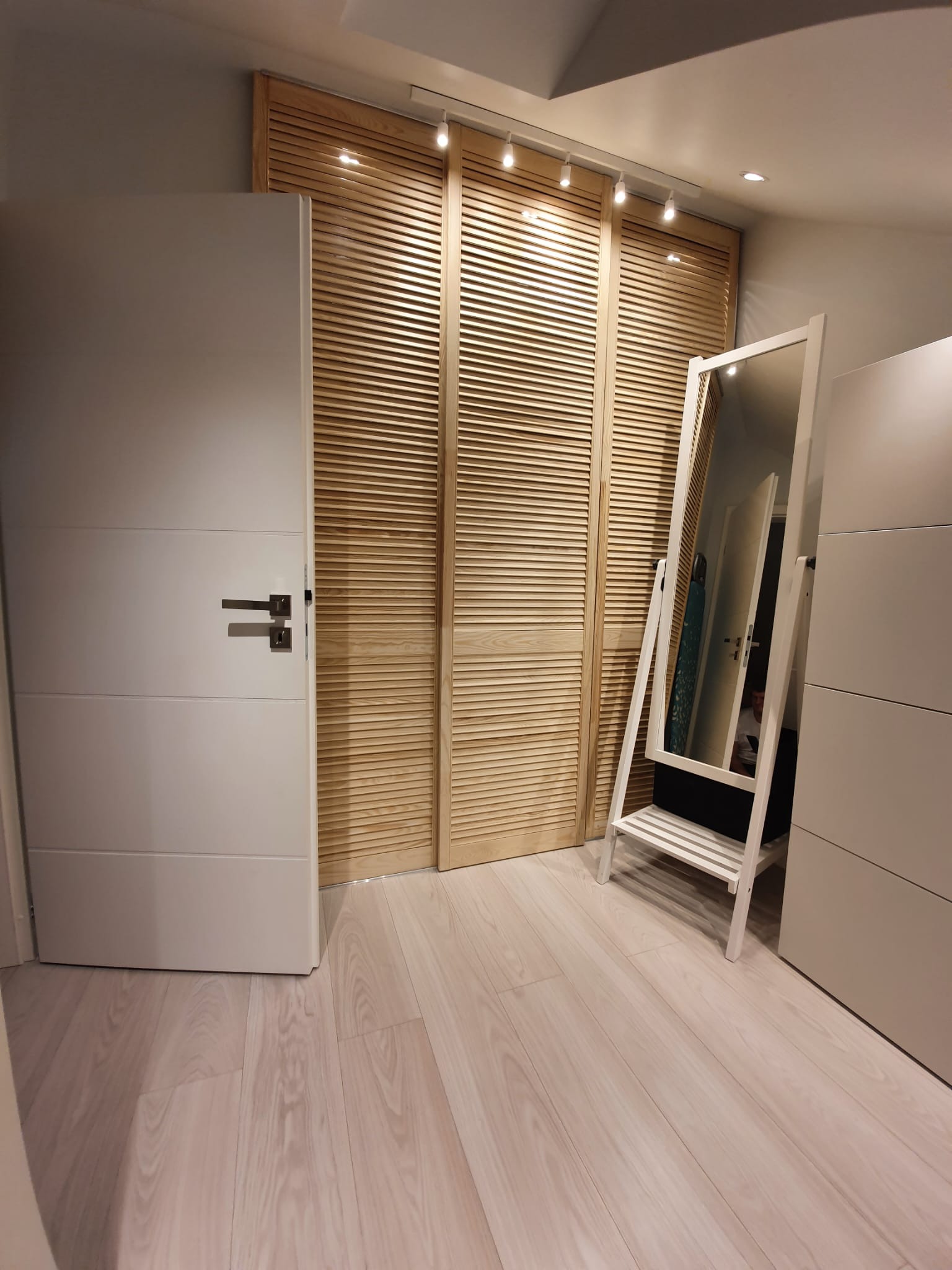Białe drzwi z poziomymi panelami i drewniane żaluzje przesuwne w tle, uzupełnione lustrem stojącym w Słupsku. Jasna podłoga i oświetlenie punktowe.