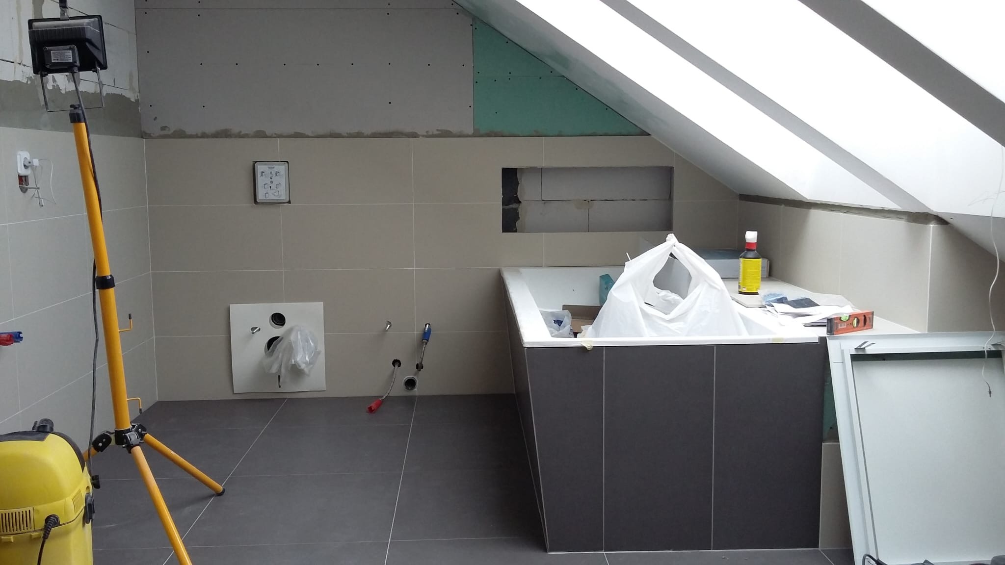 Łazienka na poddaszu w trakcie remontu: widoczne płytki, wanna obudowana ciemnymi płytkami, przygotowane ściany pod zabudowę z płyty karton-gips, oświetlenie robocze.