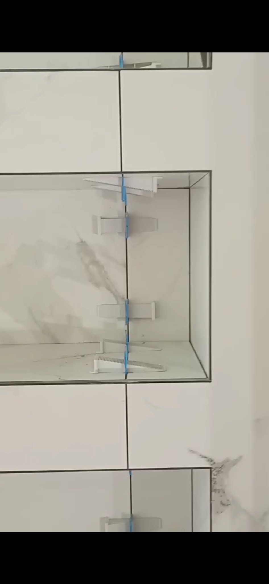 Białe płytki imitujące marmur w trakcie montażu, z użyciem niebieskich klinów poziomujących, tworzące wnękę w ścianie. Widoczne ślady po kleju.