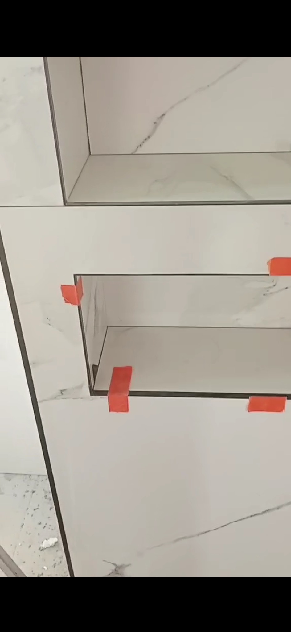 Wykończona wnęka w łazience z białych płytek imitujących marmur, zabezpieczona pomarańczowymi klinami dystansowymi. Widoczne fugi i precyzyjne cięcia.