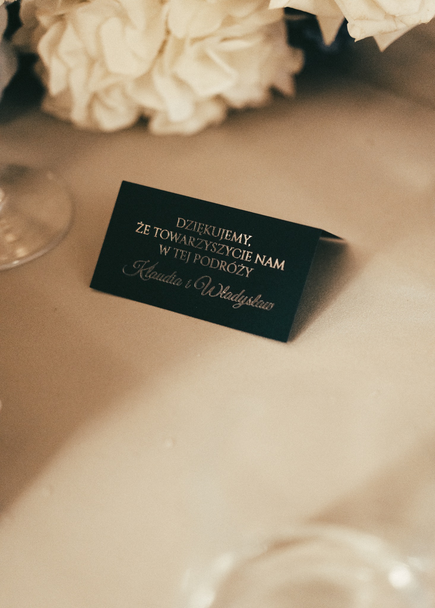 Elegancka winietka z podziękowaniem dla gości, leżąca na stole weselnym. Czarny kartonik ze złotym napisem, kwiaty w tle, subtelne oświetlenie.