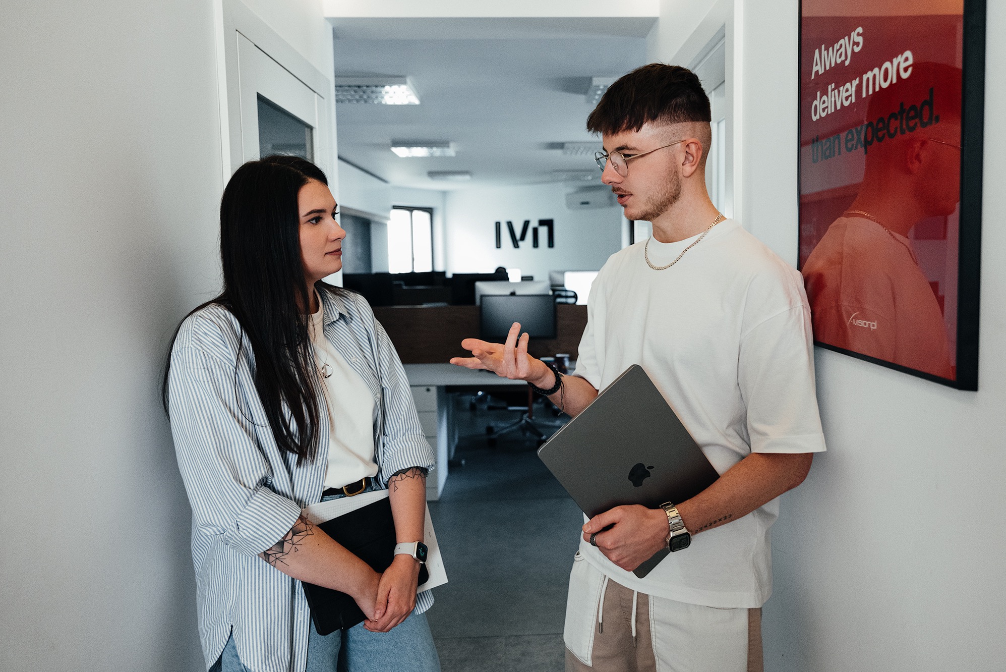Dwie osoby w biurze rozmawiają, mężczyzna trzyma laptopa. W tle widoczne logo firmy i plakat motywacyjny. Jasne wnętrze, naturalne światło.